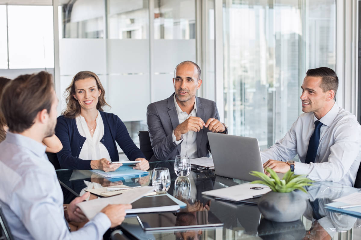 Multiethnic group of business people sitting in annual meeting in conference room. Mature leader with businessmen and businesswomen discussing. Busy partners working together in modern office.